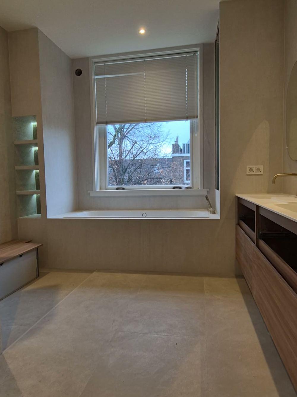 Badkamer met een ingebouwd bad onder het raam, verlichte wandnissen, een houten bankje en een houten wastafelmeubel met goudkleurige kraan in Amsterdam-Zuid.