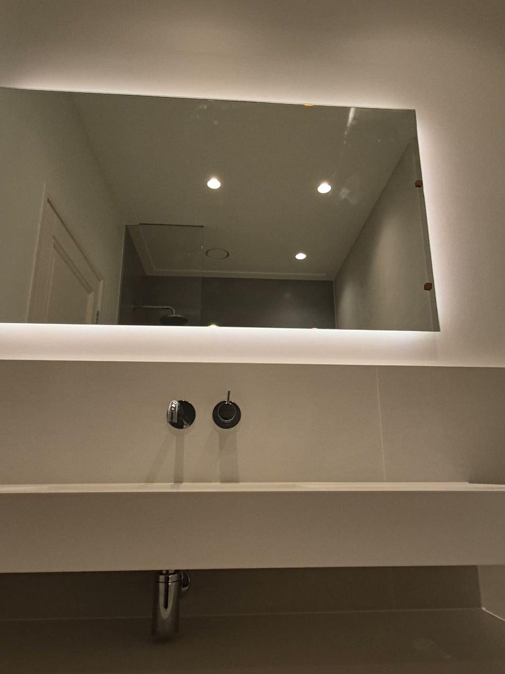 Brede witte wastafel met een verchroomde inbouwkraan en een spiegel met achtergrondverlichting in een badkamer in De Pijp.