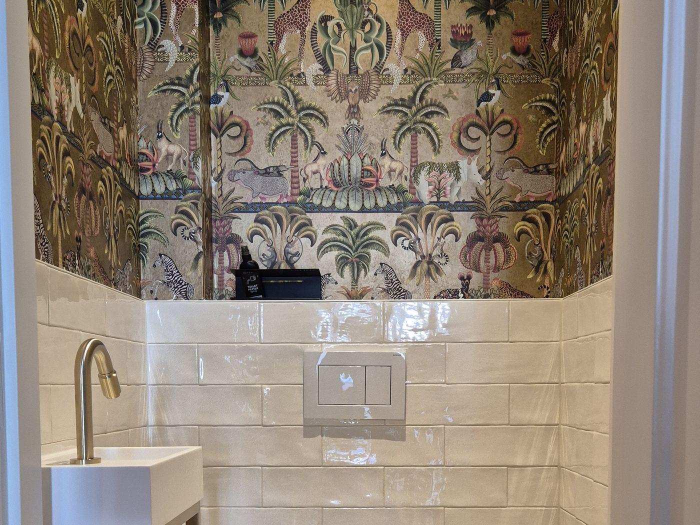 Toilet met behang met planten en dieren boven witte glanzende wandtegels, een witte bedieningsplaat en een fonteintje met goudkleurige kraan in Oud-Zuid.