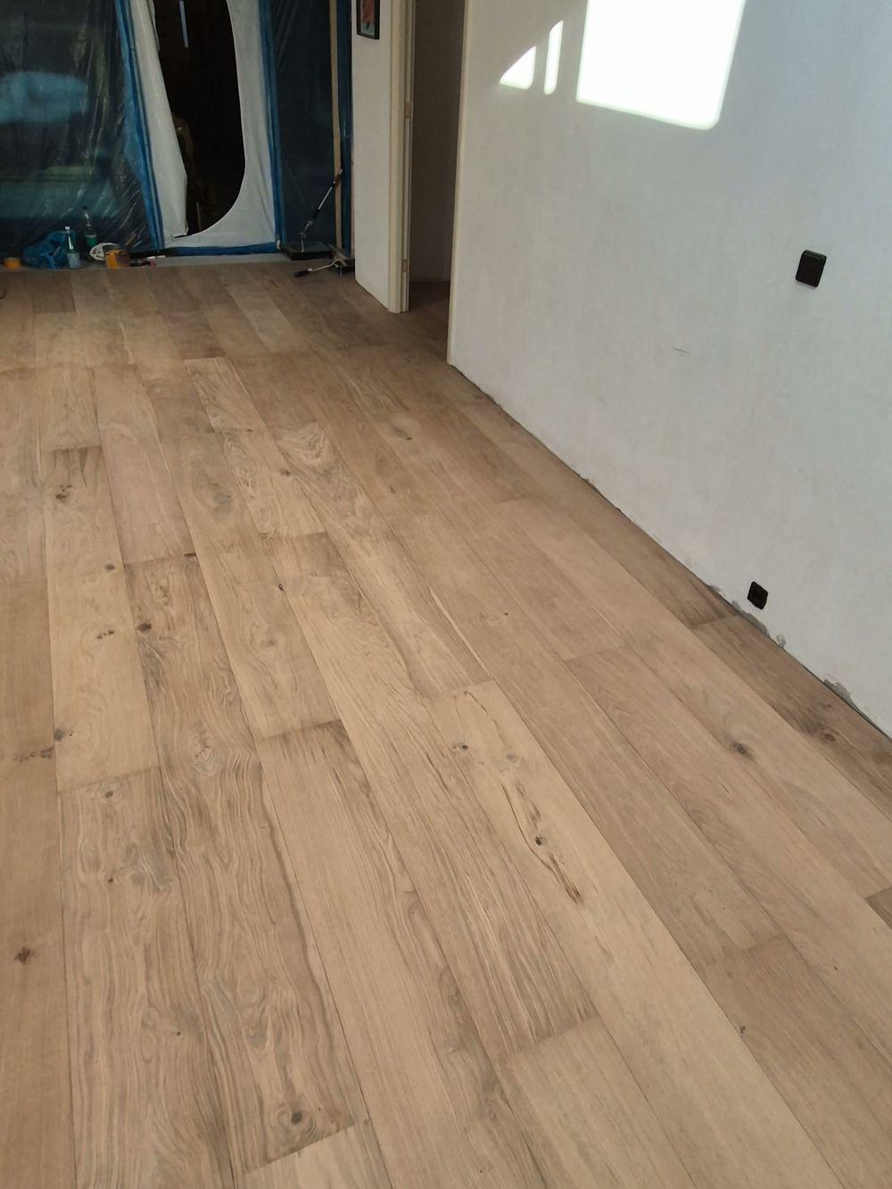 Close-up van een nieuw gelegde vloer van brede planken met zichtbare noesten en nerf tot aan een witte muur in de Jordaan.
