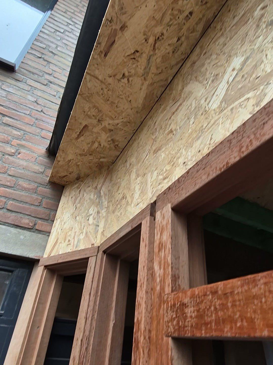 Nieuwe entreeaanbouw tegen een bakstenen gevel: hardhouten kozijnen onder een dakrand van OSB (in uitvoering).