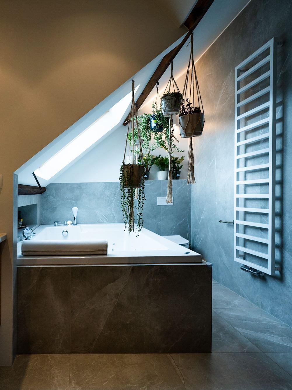 Attic bathroom with a built-in bath under a roof window, grey stone-look tiles, hanging plants and a white towel radiator.