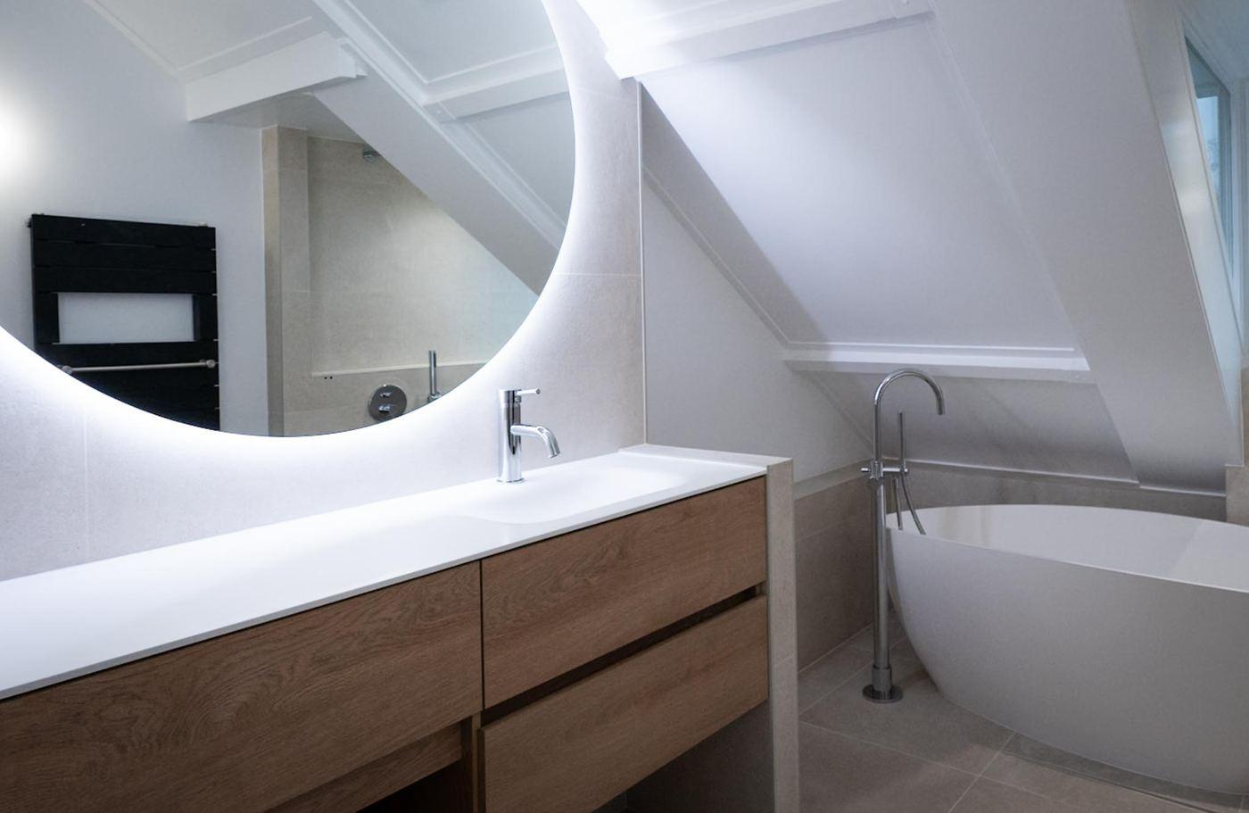 Attic bathroom with a freestanding bath under the roof slope, a wooden vanity unit and a round backlit mirror.