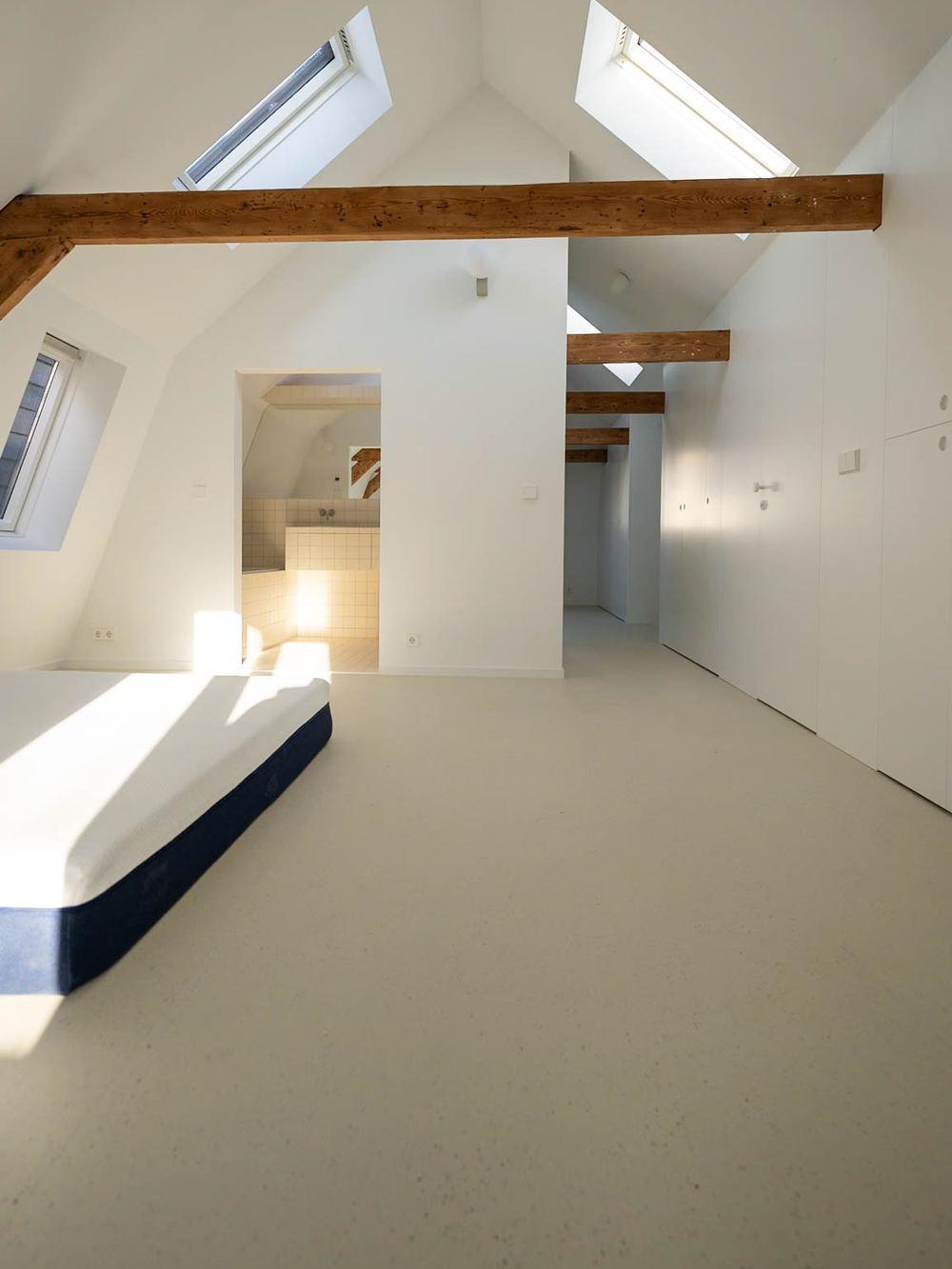 Bright attic bedroom with roof windows, exposed timber beams, white built-in wardrobes and an open doorway to a tiled bathroom.