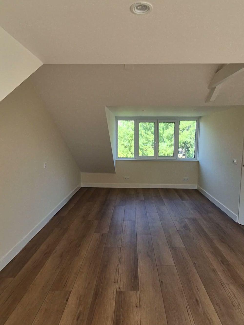 Empty attic room with a dormer window, sloping walls and a wood-look floor.